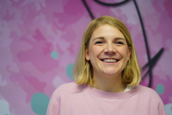 Lächelnde Frau mit blonder Haarpracht vor pinkem, gemustertem Hintergrund.